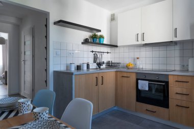 Ahşap mobilyaları, duvarlarında rafları ve beyaz fayansları olan rahat mutfağın içi. Masalı ve sandalyeli yemek odası. Şık tasarım.