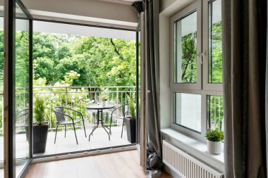 Yeni dairesinin büyük balkon penceresi yeşil manzaralı ve sandalyeli bir masa. Verandalı modern bina.