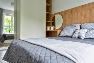 Modern yatak odasının içi rahat, gri yastıklı, pencereli, ahşap duvarlı ve aynalı. Mobilyalı ve yeni tasarımlı bir otel odası..