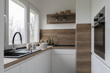 Modern mutfak iç mimarisi, pencerenin yanında lavabo ve duvarda ahşap fayanslar, şık tasarım. Küçük mutfakta beyaz mobilya ve fırın..