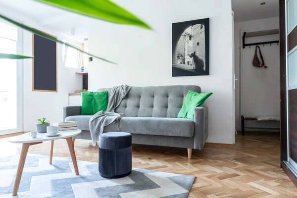 Современная гостиная с серым диваном, пуфом и столом в скандальном дизайне. Деревянный пол в светлом интерьере с изображением на белой стене. Уютный дом.