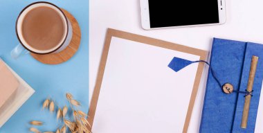 Beyaz ve mavi arkaplanda boş defter, kalem, kahve ve telefon bulunan yaratıcı ofis masası. Panoramik pankart ve üst görünüm.