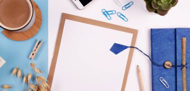 Beyaz ve mavi arkaplanda boş defter, kalem, kahve ve telefon bulunan yaratıcı ofis masası. Panoramik pankart ve üst görünüm.