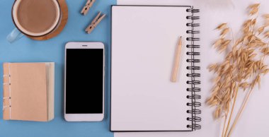 Bir fincan kahve, boş bir defter, boş ekranlı akıllı telefon ve beyaz mavi arka planda malzeme ofisi, üst manzara, günlük ofis hayatı. Panoramik pankart, üst görünüm