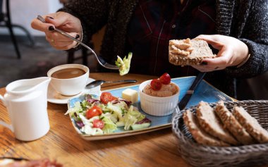 Kadın restoranda sağlıklı bir vejetaryen kahvaltısı yiyor. Sebze salatası, domatesli humus ve masada bir tabak ekmek. Kadın eli için kapatın..