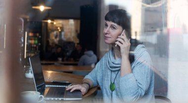 Kadın dizüstü bilgisayarda çalışıyor ve kafede bir fincan kahveyle telefonda konuşuyor. Pencerenin arkasında yansıması olan sıradan bir iş kadını. Serbest Çalışan 40 Yıl.