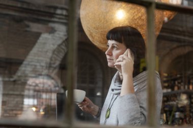 Kadın dizüstü bilgisayarda çalışıyor ve kafede bir fincan kahveyle telefonda konuşuyor. Pencerenin arkasında yansıması olan sıradan bir iş kadını. Serbest Çalışan 40 Yıl.