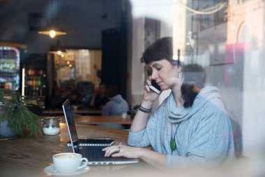 Kadın dizüstü bilgisayarda çalışıyor ve kafede bir fincan kahveyle telefonda konuşuyor. Pencerenin arkasında yansıması olan sıradan bir iş kadını. Serbest Çalışan 40 Yıl.