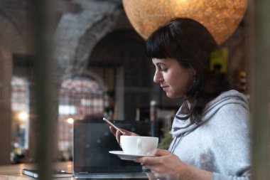 Genç bir kadın çalışırken kahve içer ve telefon kullanır. Merkez ofis. İş kadını ve penceredeki yansıma.