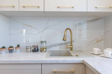 Modern bir dairede lüks bir mutfak ecru mobilyaları, duvarında mermer parlak fayanslar ve altın musluklu mutfak lavabosu. Emlak.