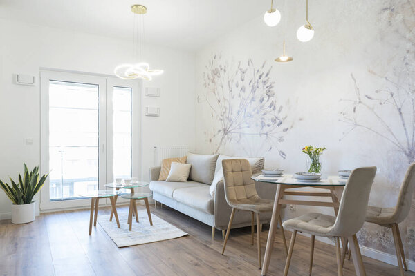 Modern living room with dining table with stylish chairs. White wall with wallpaper and luxurious interior in apartment with window. Hotel with furniture. Real estate.