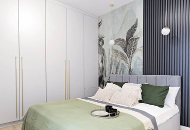 İçinde yatak örtüsü ve yatak örtüsü olan rahat bir otel odası. Duvarda yeşil yapraklarla desenli duvar kağıdı var. Şık dekorlu modern ve minimalist yatak odası. Emlak.