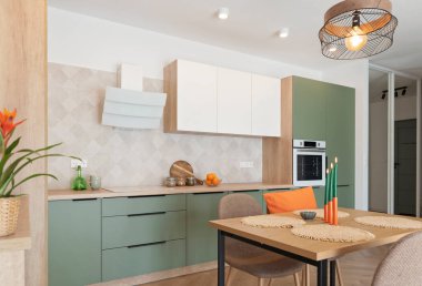 Masası ve sandalyeleri olan modern bir mutfak. Dolabı, fırını, saçakları ve turuncu süslemesi olan yeşil ve beyaz mobilyalar. Yeni dairede tasarım. Emlak.