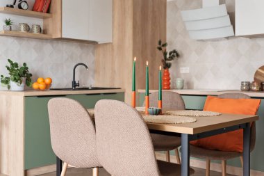 Modern mutfak ve sandalyeli ahşap yemek masası. Yeşil mobilyalar, duvarda bej fayanslar ve turuncu süslemeler. Zarif tasarımı olan yeni bir ev. Emlak.