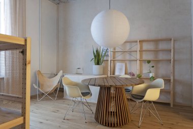 Modern bir mutfağın içinde yemek odası, tasarım koltuğu, ahşap zemin ve beton tavanda beyaz lamba olan yuvarlak bir masa. Oteldeki rahat oturma odası..