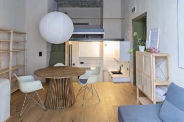 Modern bir mutfağın içinde yemek odası, tasarım koltuğu, ahşap zemin ve beton tavanda beyaz lamba olan yuvarlak bir masa. Oteldeki rahat oturma odası..