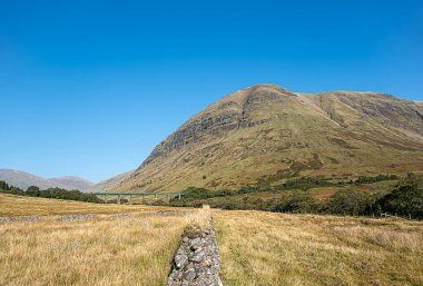 Landscape photography of mountain Beinn Mhanach and train viaduct in the valley Glen Auch, Scotland, UK, rocky wall, sunlight with blue sky, travel, hiking, hill walking clipart