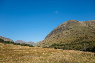 Landscape photography of mountain Beinn Mhanach and train viaduct in the valley Glen Auch, Scotland, UK, rocky wall, sunlight with blue sky, travel, hiking, hill walking clipart