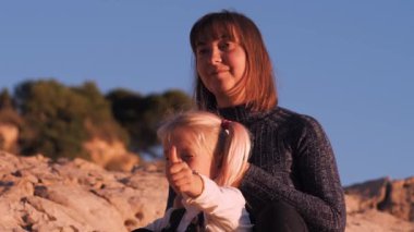 Girl with happy mother on blue sky and rocky mountain background. Child showing thumbs up. Close-up