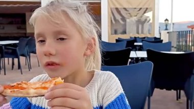 Blonde pigtail girl enjoys biting a pizza in restaurant. Close-up