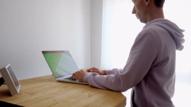 A man using laptop near window at home. Green chroma key screen. A freelancer wearing hoodie typing on notebook at standing desk. Medium shot.
