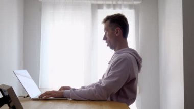 Young man using laptop near window at home. A freelancer wearing hoodie typing on notebook surfing web at standing desk. Medium shot.