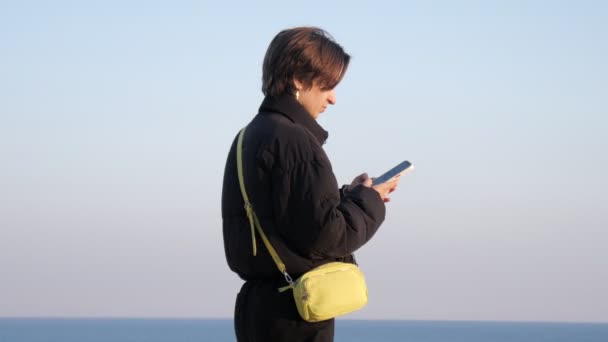 10代の少女は海岸沿いの道 青い空と海を背景にスマートフォンでチャット 自然との時間を楽しむ — ストック動画