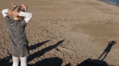 Çift kuyruklu sarışın kız kumsalda dans eder ve kumların üzerinde dans eden gölgesini seyreder. Orta boy.