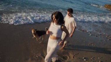 Beyaz elbiseli genç beyaz bir çift sahilde köpeklerle oynuyor. Erkek ve kadın gün doğarken sahil şeridinde koşuyorlar.