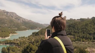 Genç kız dağ yolunda akıllı telefondan video çekiyor. Arka planda göl var. Doğanın tadını çıkarıyorum. 