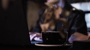 Laptop klavyesinde yazan genç bir kadının arka planında bir fincan kahve. Üç çok ırklı insan bar restoranının masasında bir projede birlikte çalışıyor.. 