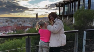 Anne ve kızının yukarıdan güzel bir şehrin tadını çıkarışının dikiz görüntüsü. Arkaplanda dağ aralığı ve bulutlu gökyüzü.