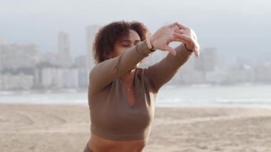 Kahverengi spor kıyafetleri içinde gülümseyen genç sporcu kadın, arka planda deniz ve gökdelenlerle kumsalda bir dizi esneme egzersizi yapıyor..