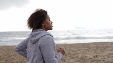 Sahilde ağır çekimde koşan kapüşonlu genç bir kadın. Arka planda deniz dalgaları var. Sağlıklı bir yaşam tarzının tadını çıkarıyor. Orta boy.