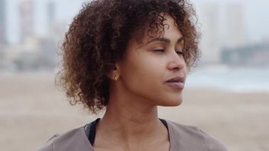 Kıvırcık saçlarını sallayıp kumsalda duran, güneşli yaz gününde manzaralı okyanusa ve dağ manzarasına bakan mutlu İspanyol kadının yakın çekimi.