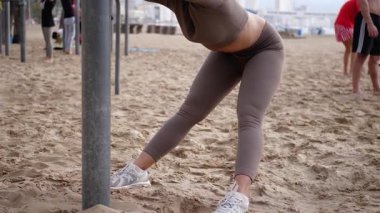 Açık hava fitness antrenmanı sırasında kumlu plajda kollarını arkaya uzatan kıvırcık saçlı genç atletik kadın esneklik ve sağlığını gösteriyor.