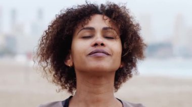 Mutlu, kıvırcık saçlı, kumlu sahilde duran, güneşli yaz gününde manzaralı okyanusa ve dağ manzarasına bakan İspanyol kadının yakın çekimi.