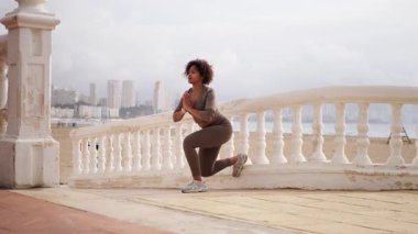 Fitness modeli güneşli sahil terasında tek bacaklı çömelme alıştırması yapıyor, çarpıcı şehir manzarasına karşı denge ve güç gösteriyor..