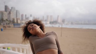 Genç İspanyol kadın plajdaki antrenmanına ara veriyor, geriye yaslanıyor ve derin derin nefes alıyor, temiz havanın ve çarpıcı şehir manzarasının tadını çıkarıyor.. 
