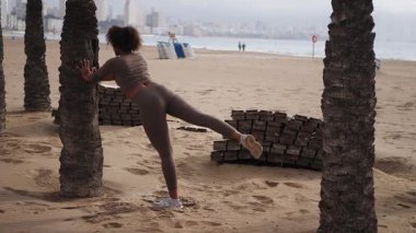 Spor kıyafetleri giyen genç İspanyol kadın sahilde palmiye ağacının yanında ayağını geri çekiyor. Arka planda deniz ve şehir var.