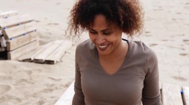 Spor kıyafetleri giymiş gülümseyen İspanyol kadın kumlu plajda bacaklarını esnetiyor. Orta boy.