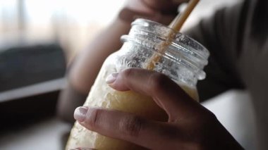 Sarı-yeşil smoothie ile bardağı tutan kadın ellerinin yakın çekimi. Genç Afrikalı Amerikalı kadın sahildeki barda otururken ferahlatıcı bir içki yudumluyor..