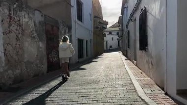 Genç bir kadın güvenli bir şekilde scooter 'la dar kaldırım taşlarında geziniyor, güneşli yolculukta canlı şehir hareketliliğini ve çocukluk özgürlüğünü yakalıyor.