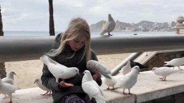 Beyaz güvercinleri beslerken beyaz bir kız neşeli anlar yaşıyor. Bir güvercin kızların kafasına oturur. Arka planda palmiye ağaçları ve tekneleri olan deniz manzaralı bir gezinti.