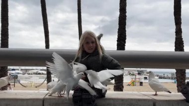 Beyaz güvercinleri beslerken beyaz bir kız neşeli anlar yaşıyor. Kızların omzunda bir güvercin oturuyor. Arka planda palmiye ağaçları ve tekneleri olan deniz manzaralı bir gezinti.