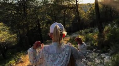 Sarışın dişi, altın gün batımında orman yolunda yürürken sırt çantalarını düzeltiyor. Ağaçlar ve sıcak akşam ışığı ile çevrili.