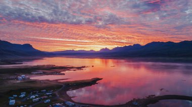 Sakin bir İzlanda fiyordunun üzerinde canlı bir günbatımı turuncu, pembe ve mor renkleri yansıtır. Siluetli dağlar ve küçük bir yerleşim yeri görünür..