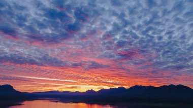 İzlanda 'da canlı bir gün batımı turuncu, pembe ve mor renkler sakin suya yansıyor, engebeli dağ siluetleri ve dramatik bulutlarla çerçevelenmiş..