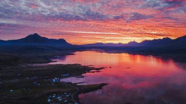 Gün batımında İzlanda fiyordunun durgun suları, engebeli zirveleri ve küçük kıyı şeridi yerleşimlerini yansıtan pembe ve turuncu renkleriyle sakin bir hava manzarası..
