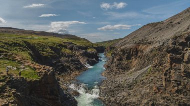 Yeşil bitki örtüsü, kayalık uçurumlar ve küçük bir şelale ile İzlanda 'daki engebeli bir kanyondan canlı bir mavi nehir akar..